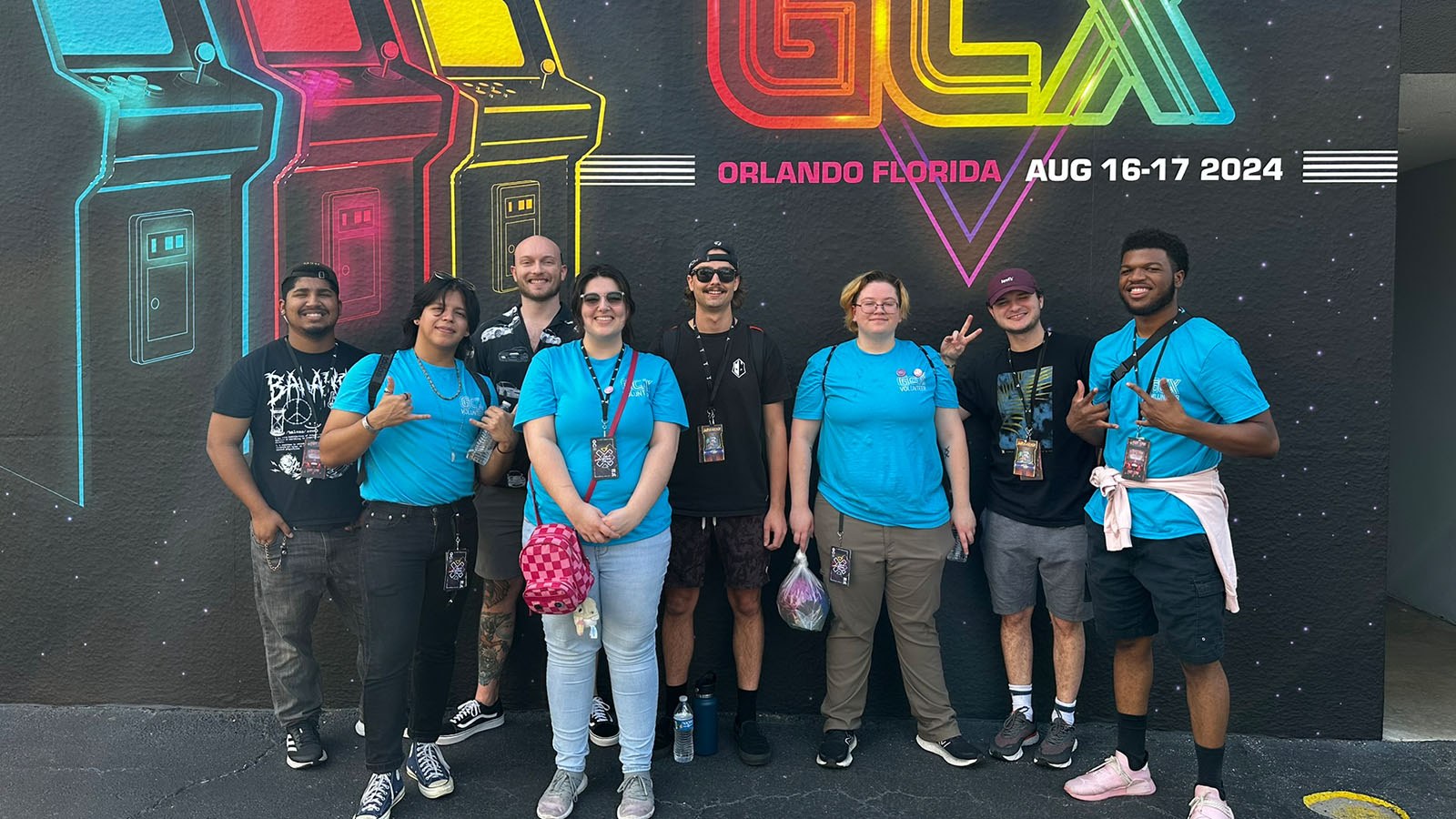 A group of eight people pose in front of a mural that reads "GCX Orlando, Florida - Aug 16-17 2024," with colorful arcade designs.