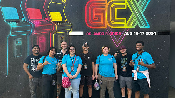 A group of eight people pose in front of a mural that reads "GCX Orlando, Florida - Aug 16-17 2024," with colorful arcade designs.