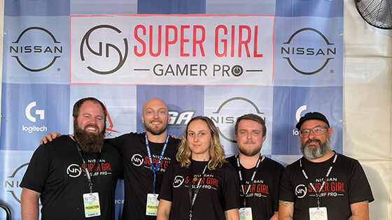 Five people wearing black shirts and badges that read "production" while standing in front of a step and repeat featuring the Super Girl Gamer pro logo and event sponsor logos.