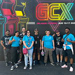 A group of eight people pose in front of a mural that reads "GCX Orlando, Florida - Aug 16-17 2024," with colorful arcade designs.